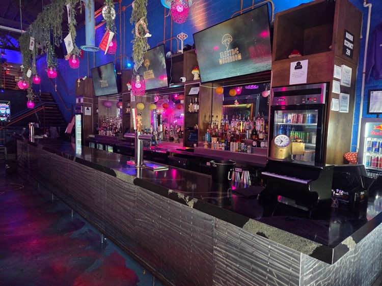 Long Electric Mushroom bar with back bar bottles and screens in pink and blue light